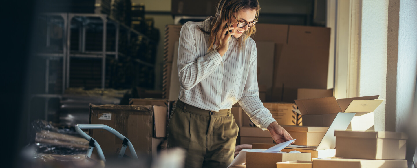 Woman surrounded by boxes sorts through paperwork while on the phone.